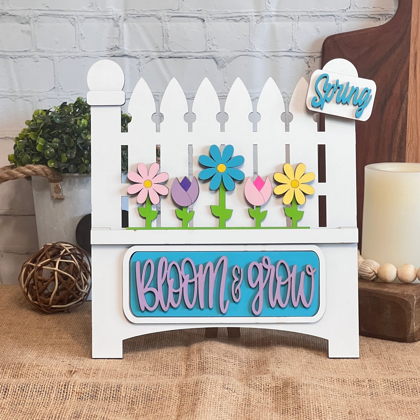 Janet's Craft Corner presents the "Bloom & Grow Interchangeable Insert" kit, featuring decorative signs with a white picket fence, colorful flowers, and "Bloom & Grow" in blue and pink. Includes a tag reading "Spring Blooms," ideal for adding charm to any setting alongside bricks, lush plants, a woven ball, or candle.