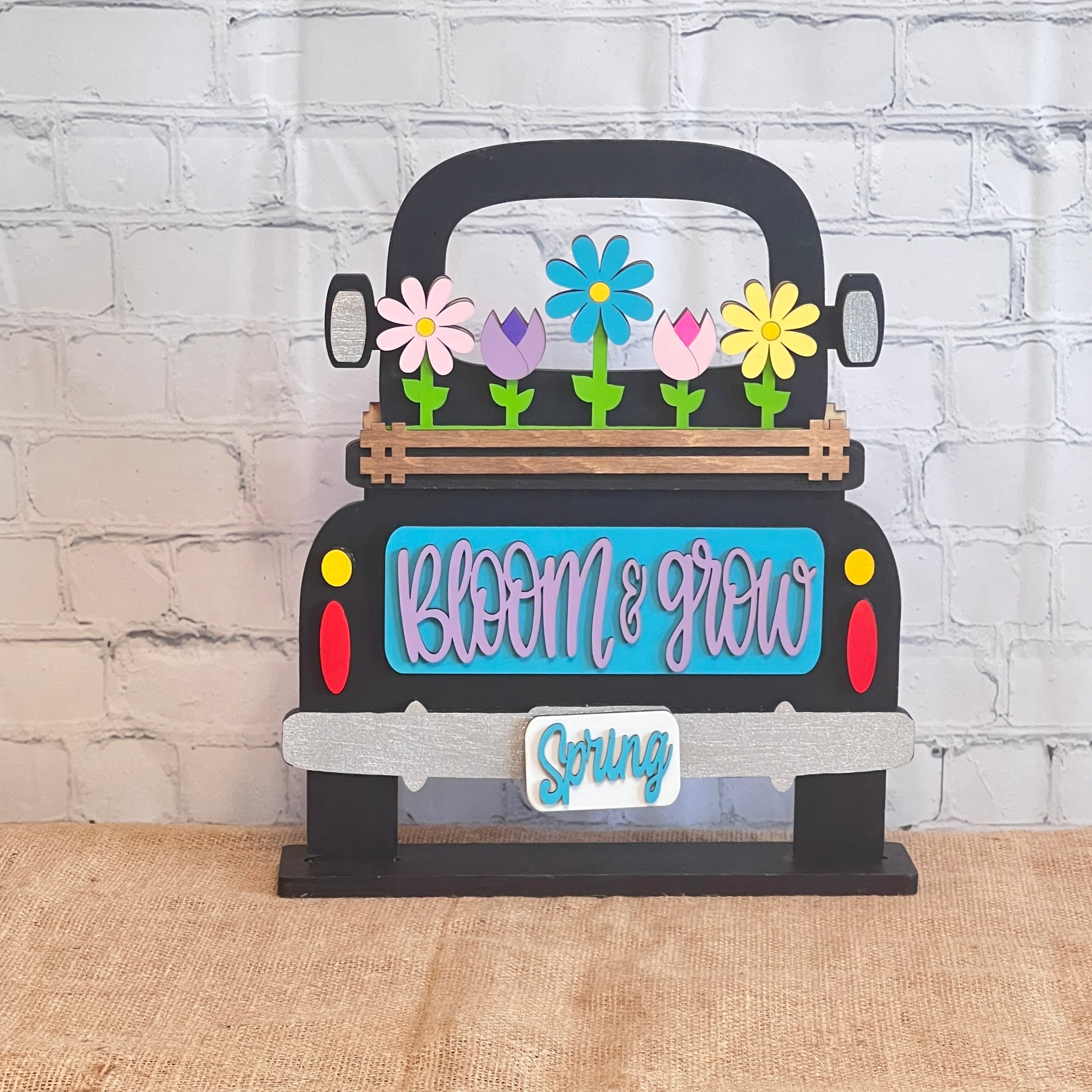 This antique wooden truck display from Janet's Craft Corner features a blue area with "Bloom & Grow" in pink, colorful Spring Blooms in the bed, "Spring" on the bumper, all set on burlap against a brick-patterned backdrop as part of the Bloom & Grow Interchangeable Insert DIY kit.