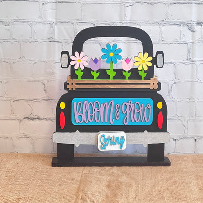 This antique wooden truck display from Janet's Craft Corner features a blue area with "Bloom & Grow" in pink, colorful Spring Blooms in the bed, "Spring" on the bumper, all set on burlap against a brick-patterned backdrop as part of the Bloom & Grow Interchangeable Insert DIY kit.