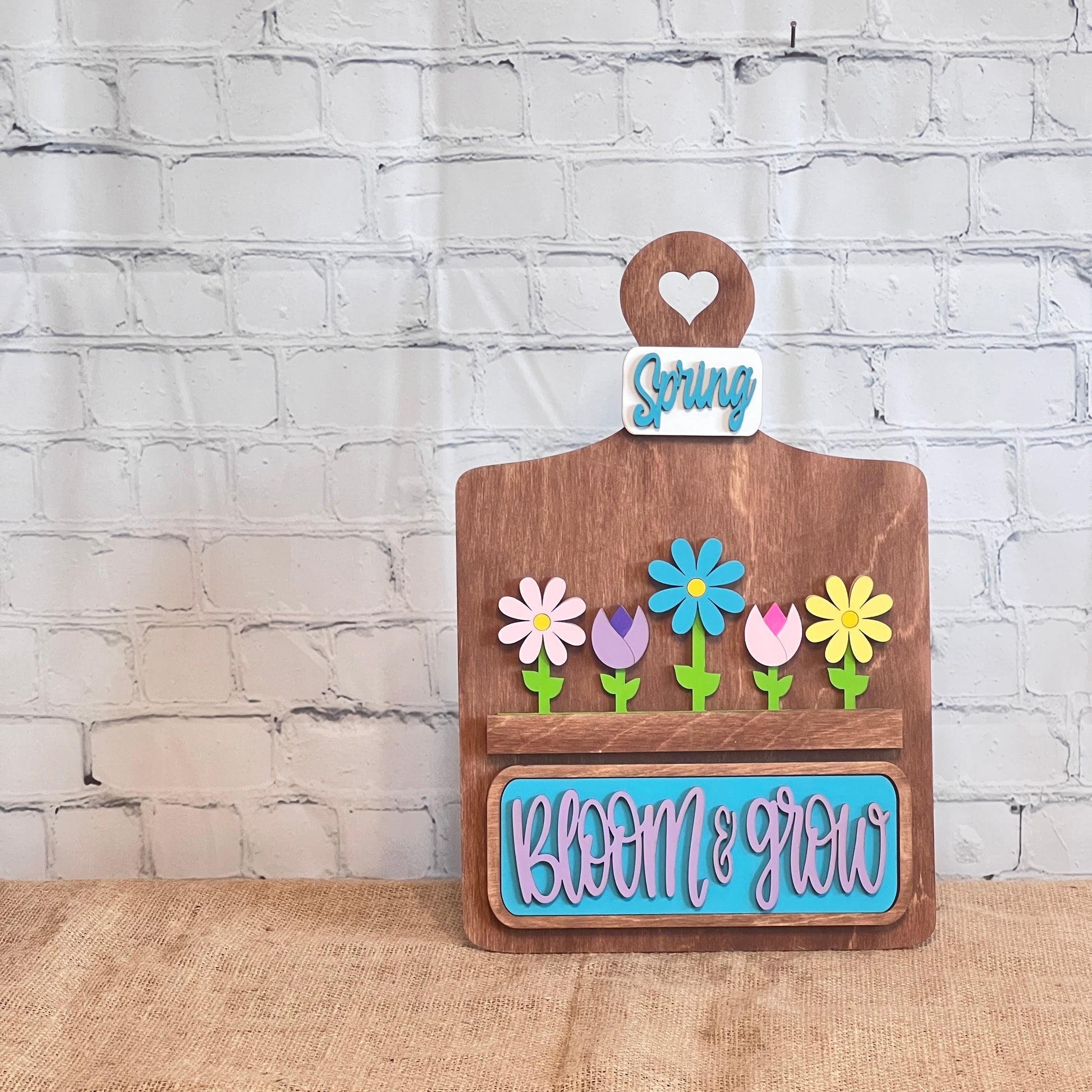 The Bloom & Grow Interchangeable Insert kit from Janet's Craft Corner, featuring colorful flowers and "Bloom & Grow" in blue on brown, complemented by a charming "Spring Blooms" insert against a white brick wall, adds a decorative touch with its 3-piece DIY home decor craft set.