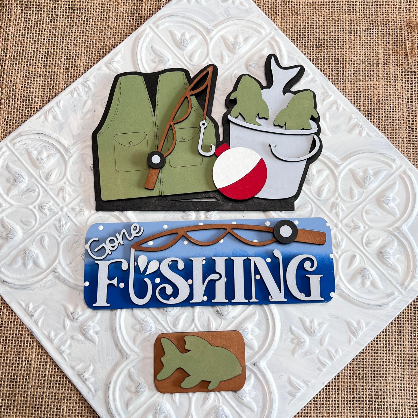 The Gone Fishing Interchangeable Insert DIY craft kit by Janet's Craft Corner includes 3 charming pieces—a green vest, fish bucket, and “Gone Fishing” sign—perfect for seasonal home decor.