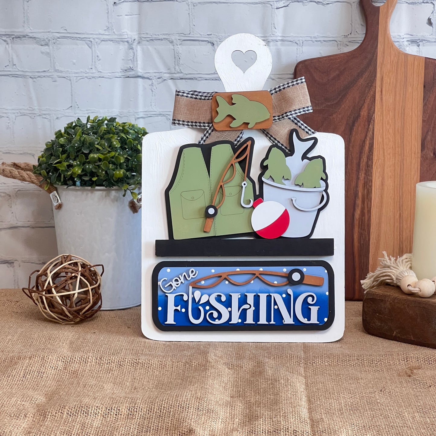 The Gone Fishing Interchangeable Insert from Janet's Craft Corner is a DIY seasonal home decor kit with 3 wood cut-out pieces: a green vest, fishing rod, and bobber over a "Gone Fishing" sign—ideal for craft enthusiasts.