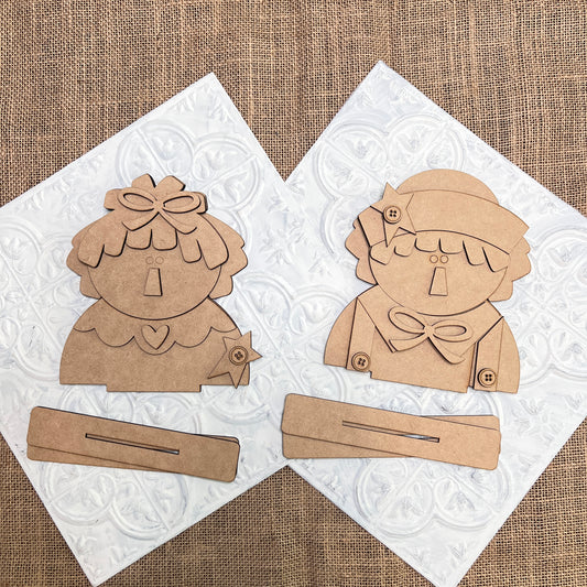 Janet's Craft Corner Raggedy Ann & Andy Shelf Sitters DIY kit includes two unfinished wooden cutouts with bases—one doll has a bow and collar, the other a hat and scarf—perfect for creating charming seasonal home decor. Set of 1.