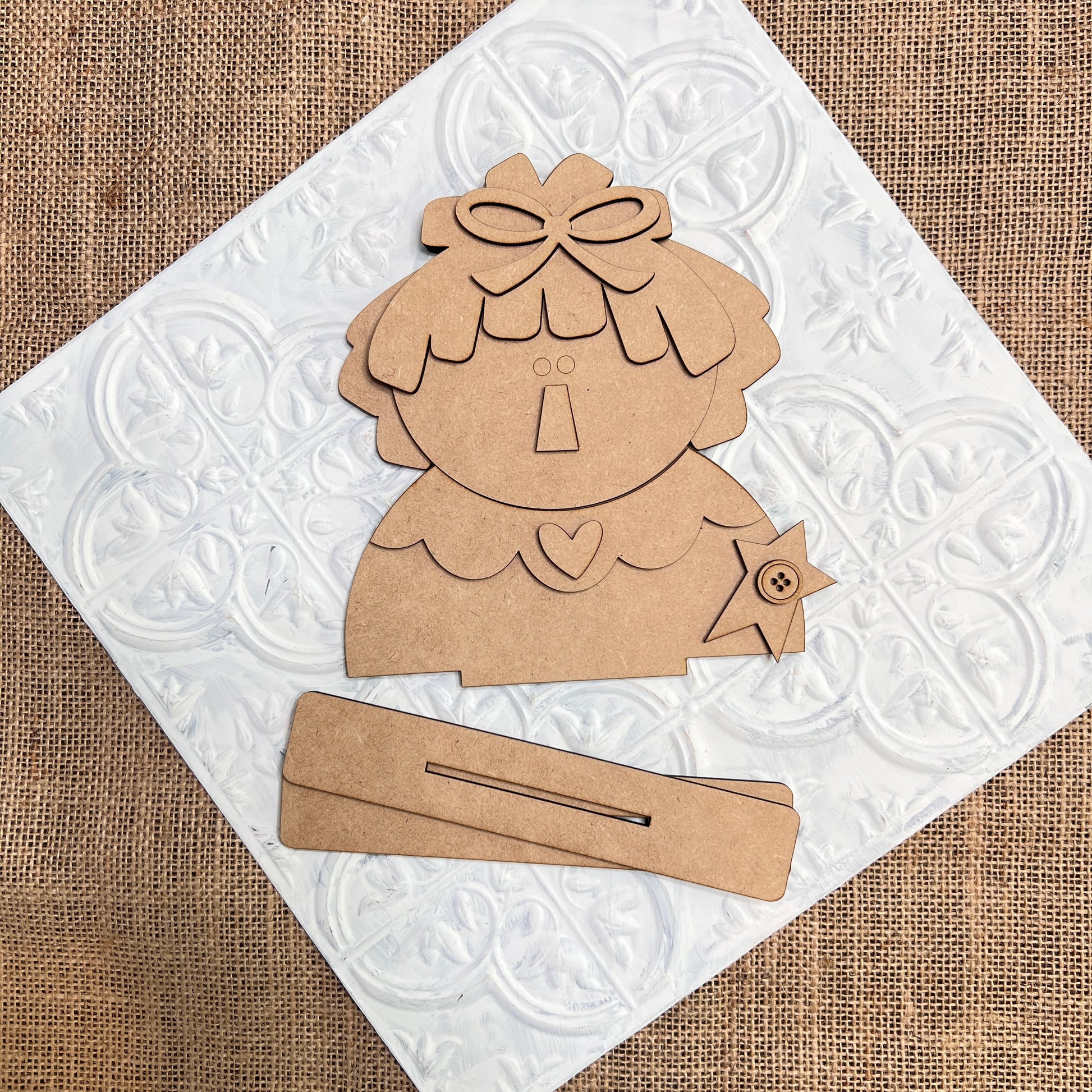 Janet's Craft Corner’s Raggedy Ann & Andy Shelf Sitters DIY kit features whimsical cardboard pieces with bow, heart, and star accents, displayed on white tile over burlap for vintage charm in your seasonal home decor creations.