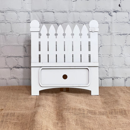 The Garden Fence Interchangeable Base - Painted from Janet's Craft Corner is a small white drawer unit featuring a picket fence craft kit on top, set against a white brick wall and brown burlap—ideal for customizable DIY home decor.