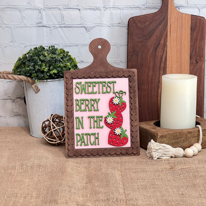 Janet's Craft Corner Berry Sweet Switchable Farmhouse Signs—Summer DIY Craft Kit, Choice of 3—features strawberries and daisies with "Sweetest Berry in the Patch" text, shown with summer décor on burlap against a white brick wall.