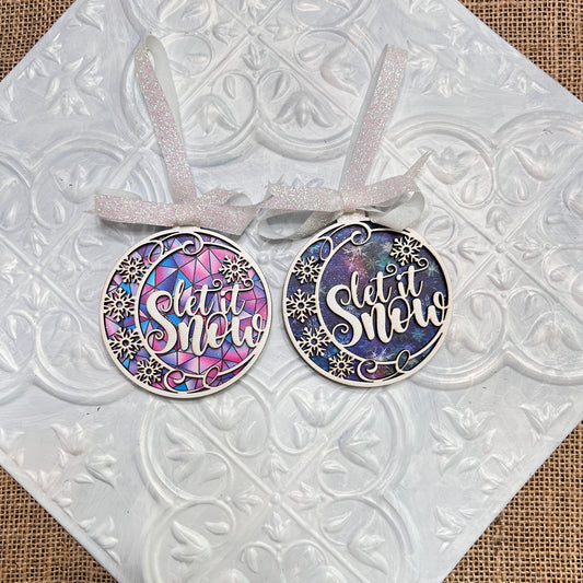 Janet's Craft Corner Let It Snow Ornaments: Two handmade round ornaments, one blue and one pink-blue stained glass, both with "Let It Snow" and snowflakes. Each includes a white ribbon and is displayed on textured white tile.