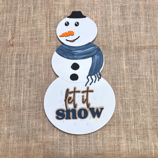The Reversible Snowman/Pumpkin Shelf Leaner from Janet's Craft Corner is a DIY seasonal home decor craft kit. Featuring a snowman with a black hat, blue scarf, carrot nose, and “let it snow” text, it's perfect for festive decorating.