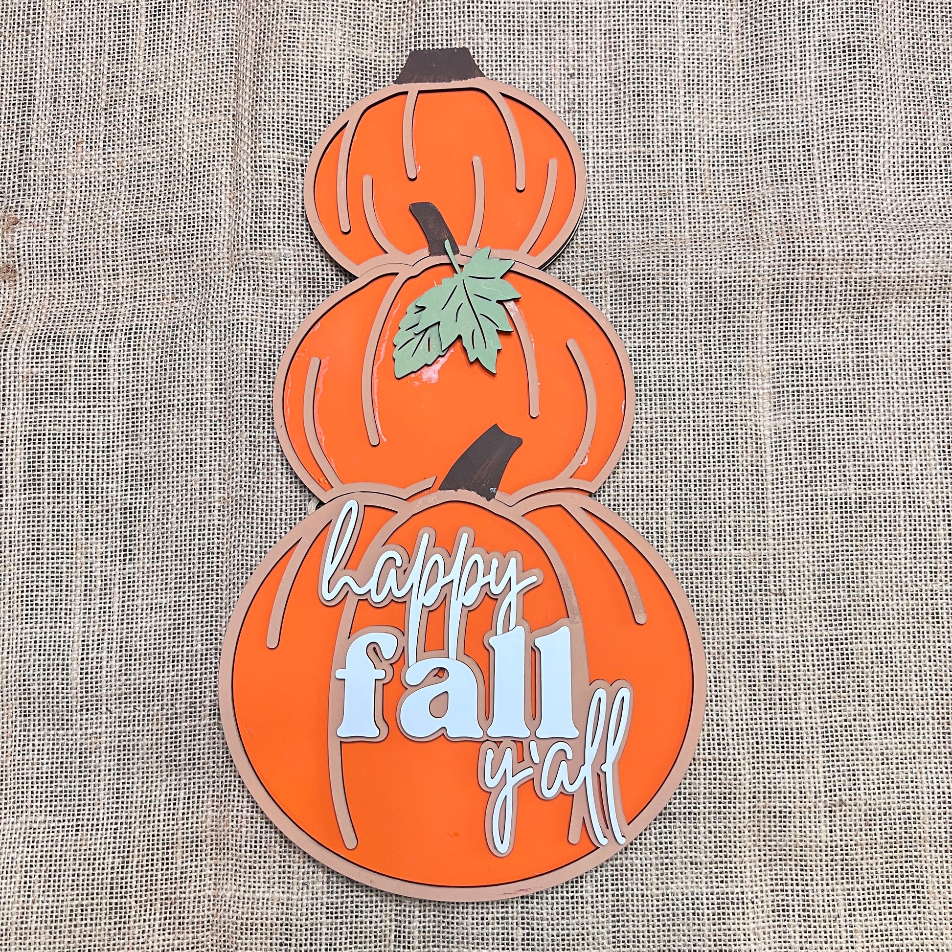 The Reversible Snowman/Pumpkin Shelf Leaner by Janet's Craft Corner is a DIY seasonal home decor kit featuring three stacked orange pumpkins on burlap; the bottom pumpkin reads "happy fall y'all," and a green leaf accents the center.