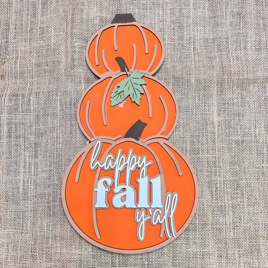 The Reversible Snowman/Pumpkin Shelf Leaner by Janet's Craft Corner is a DIY seasonal home decor kit featuring three stacked orange pumpkins on burlap; the bottom pumpkin reads "happy fall y'all," and a green leaf accents the center.