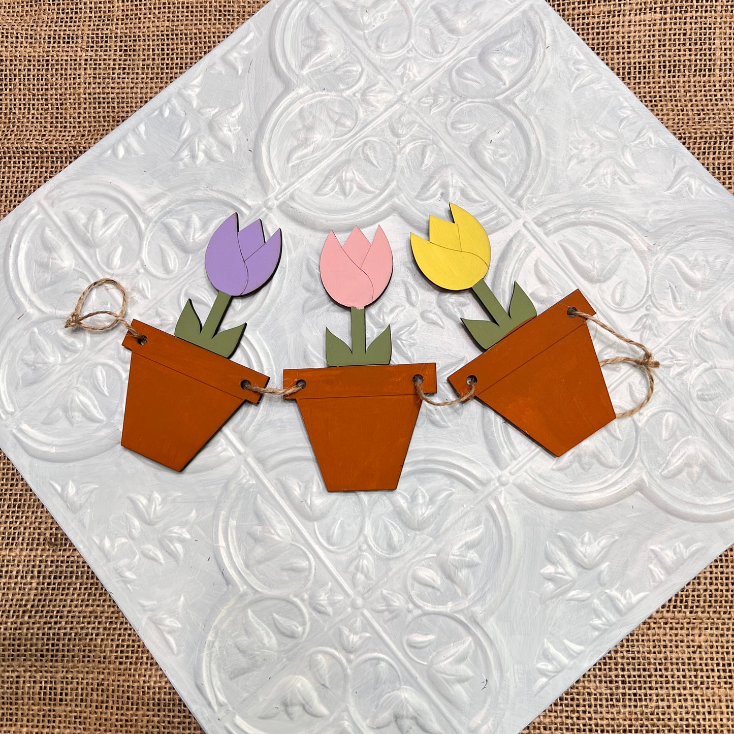 Janet's Craft Corner Tiered Tray Garland features three wooden flower pots with purple, pink, and yellow tulip shapes, displayed on textured white tile and a burlap background.