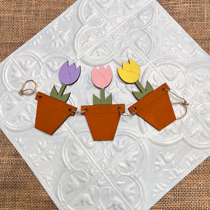 Janet's Craft Corner Tiered Tray Garland features three wooden flower pots with purple, pink, and yellow tulip shapes, displayed on textured white tile and a burlap background.