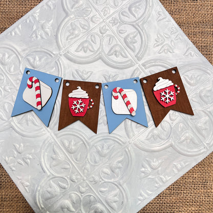 Janet's Craft Corner Tiered Tray Garlands: A festive banner with four pennants featuring red mugs of cocoa or candy canes on blue and brown backgrounds, displayed on a textured white tile.