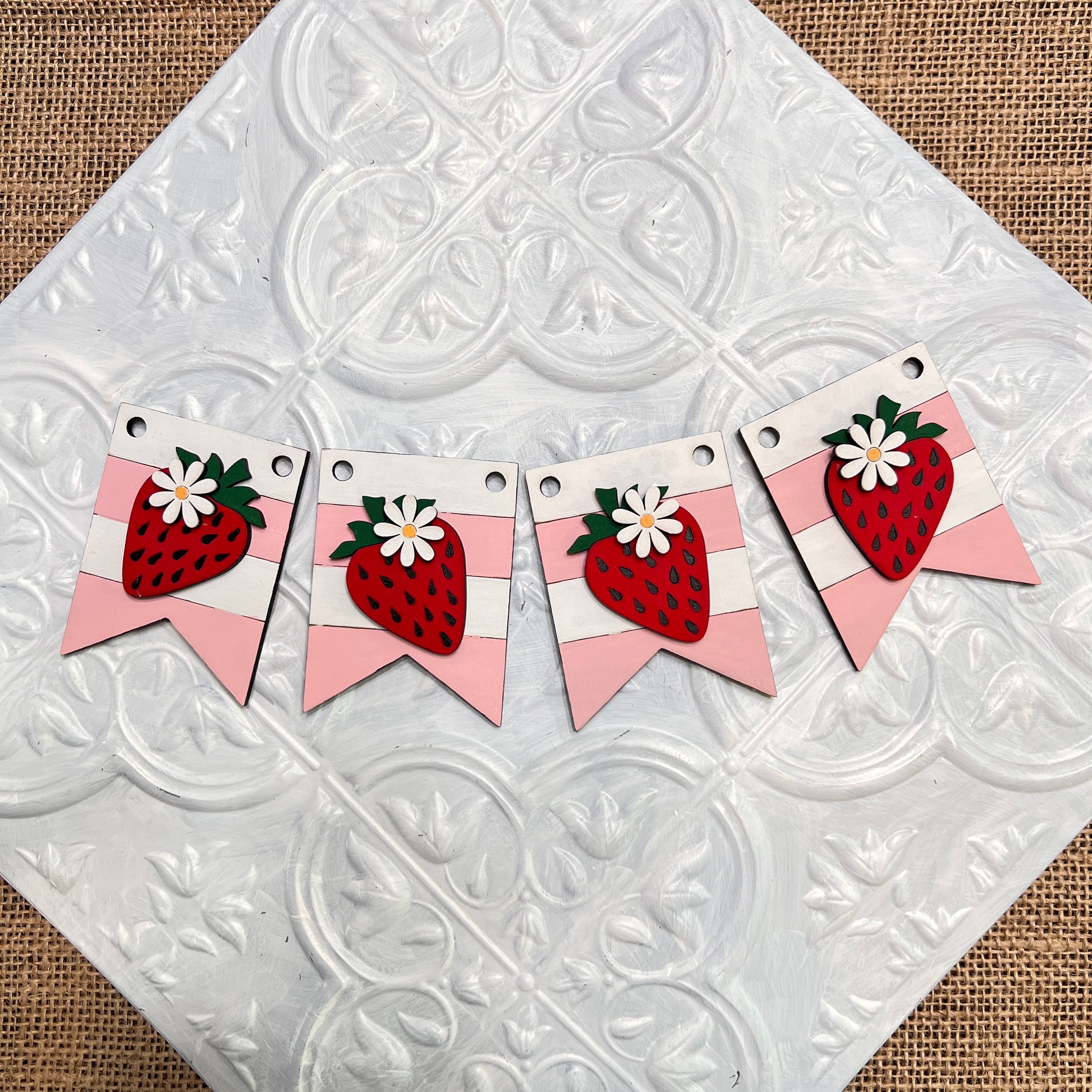 Janet's Craft Corner Tiered Tray Garlands feature four banners with pink stripes and red strawberries topped with white flowers, arranged in a row on a white embossed tile background over burlap.
