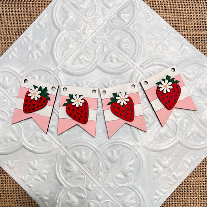 Janet's Craft Corner Tiered Tray Garlands feature four banners with pink stripes and red strawberries topped with white flowers, arranged in a row on a white embossed tile background over burlap.
