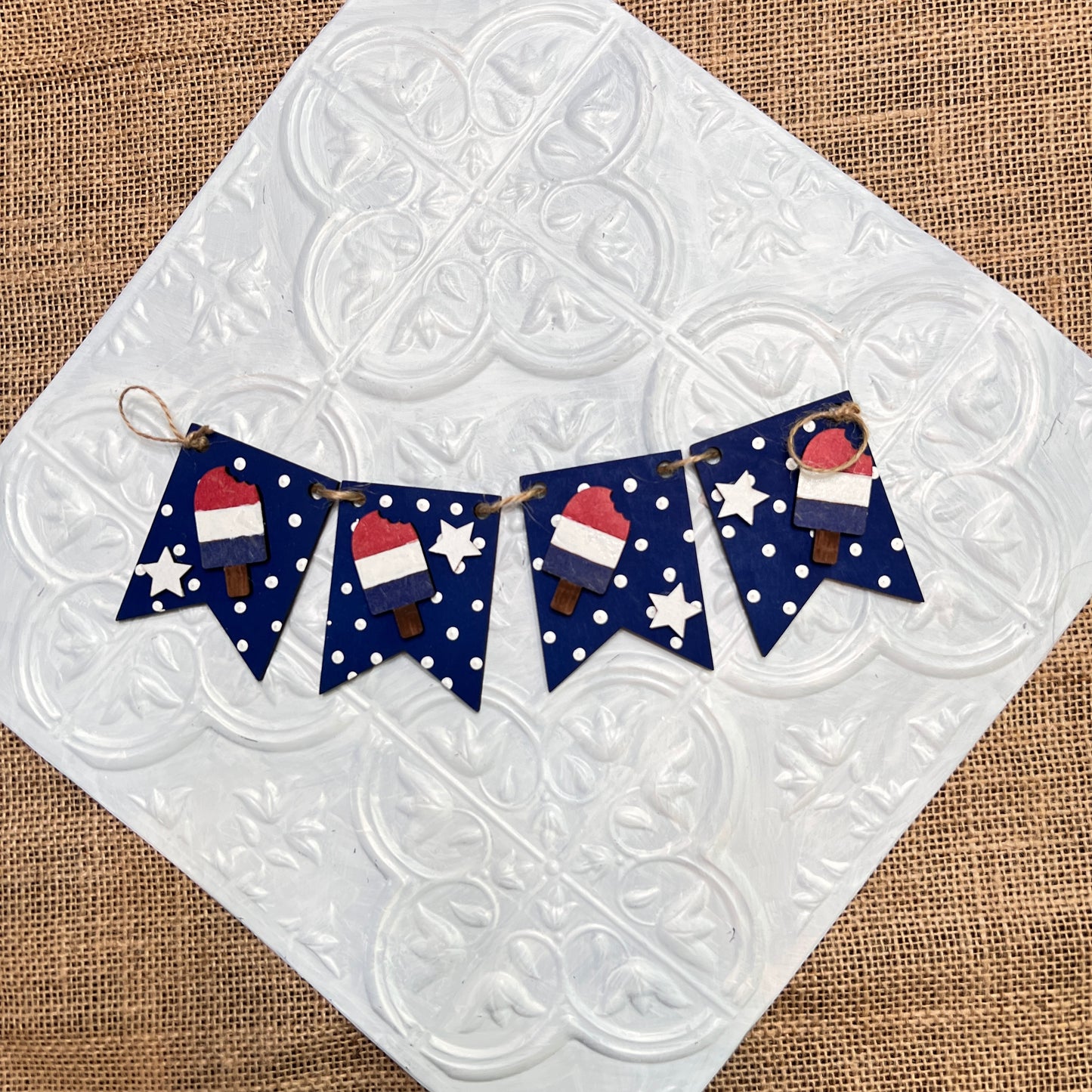 The Heartland Tiered Tray Garlands - Painted - Choice of 12 features a small banner with four dark blue pennants adorned with white dots, felt popsicles, stars, and Santa hats displayed on a textured tile and burlap background.