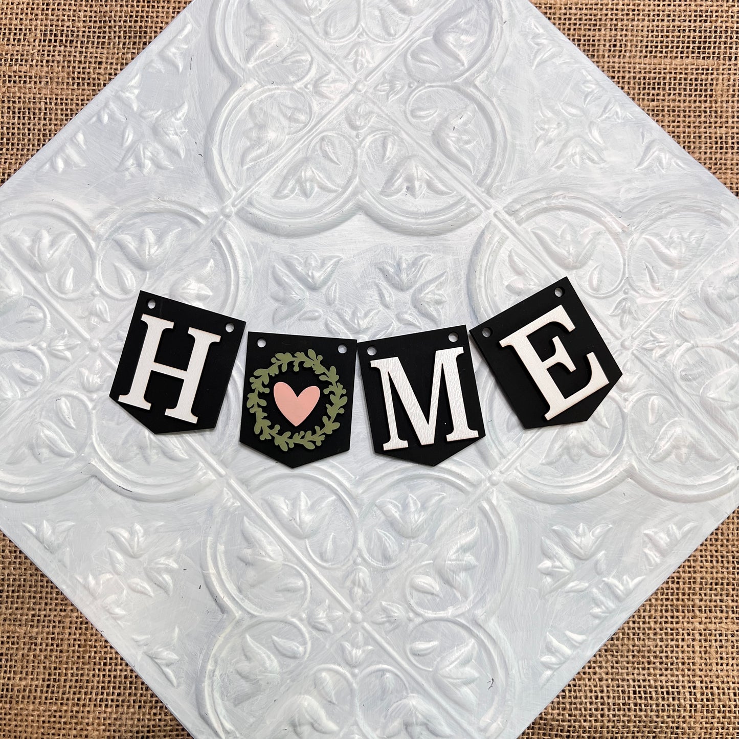 The Heartland Tiered Tray Garlands – Painted (choice of 12) feature "HOME" spelled with black pennants, a green leaf wreath and pink heart as the "O," set on a textured white and silver tile over burlap.