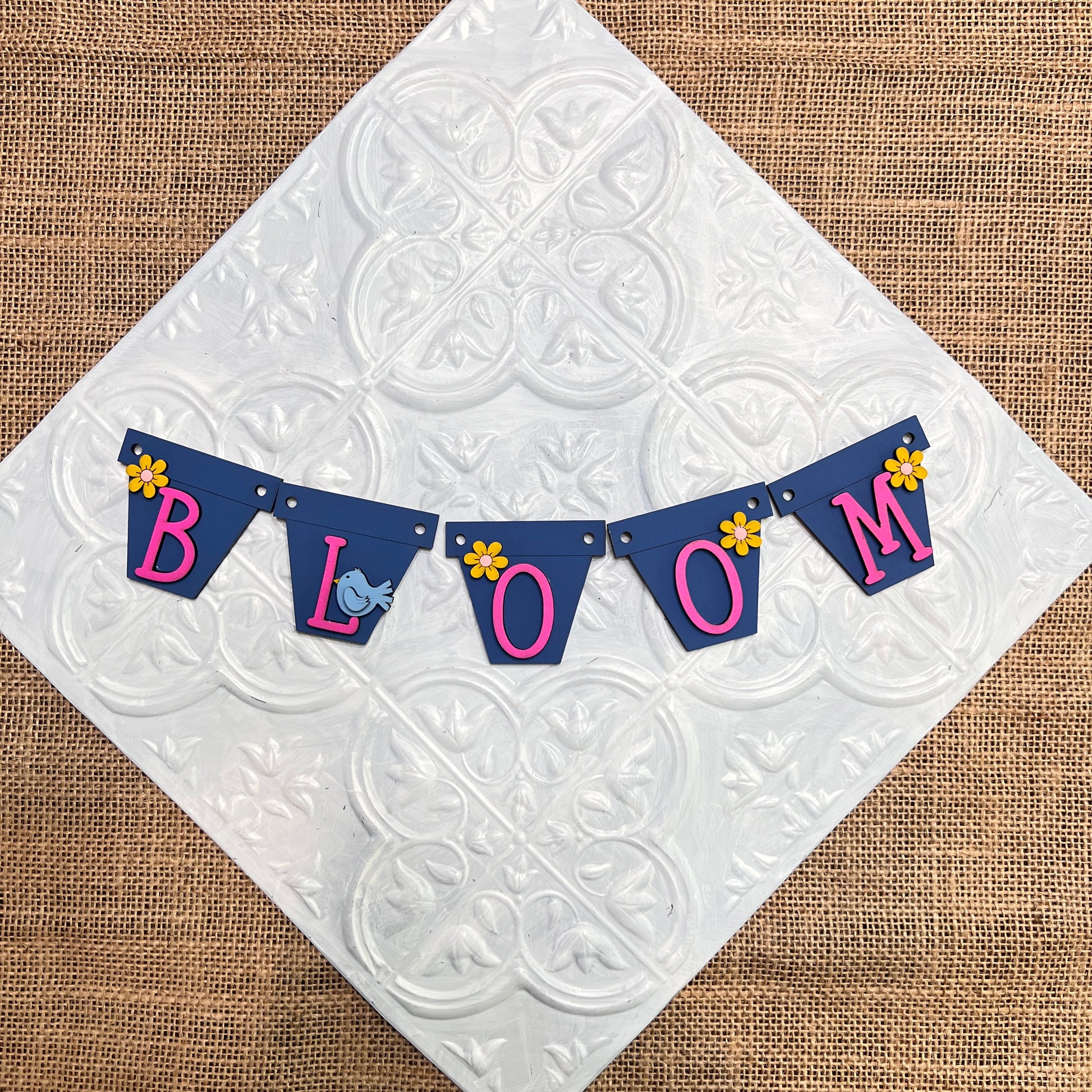 Heartland's Tiered Tray Garlands - Painted (choose from 12 styles) features a blue "BLOOM" pennant banner with pink letters, yellow flower accents, and a small blue bird, displayed on a white tile surface over burlap.