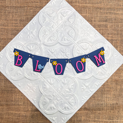 Heartland's Tiered Tray Garlands - Painted (choose from 12 styles) features a blue "BLOOM" pennant banner with pink letters, yellow flower accents, and a small blue bird, displayed on a white tile surface over burlap.