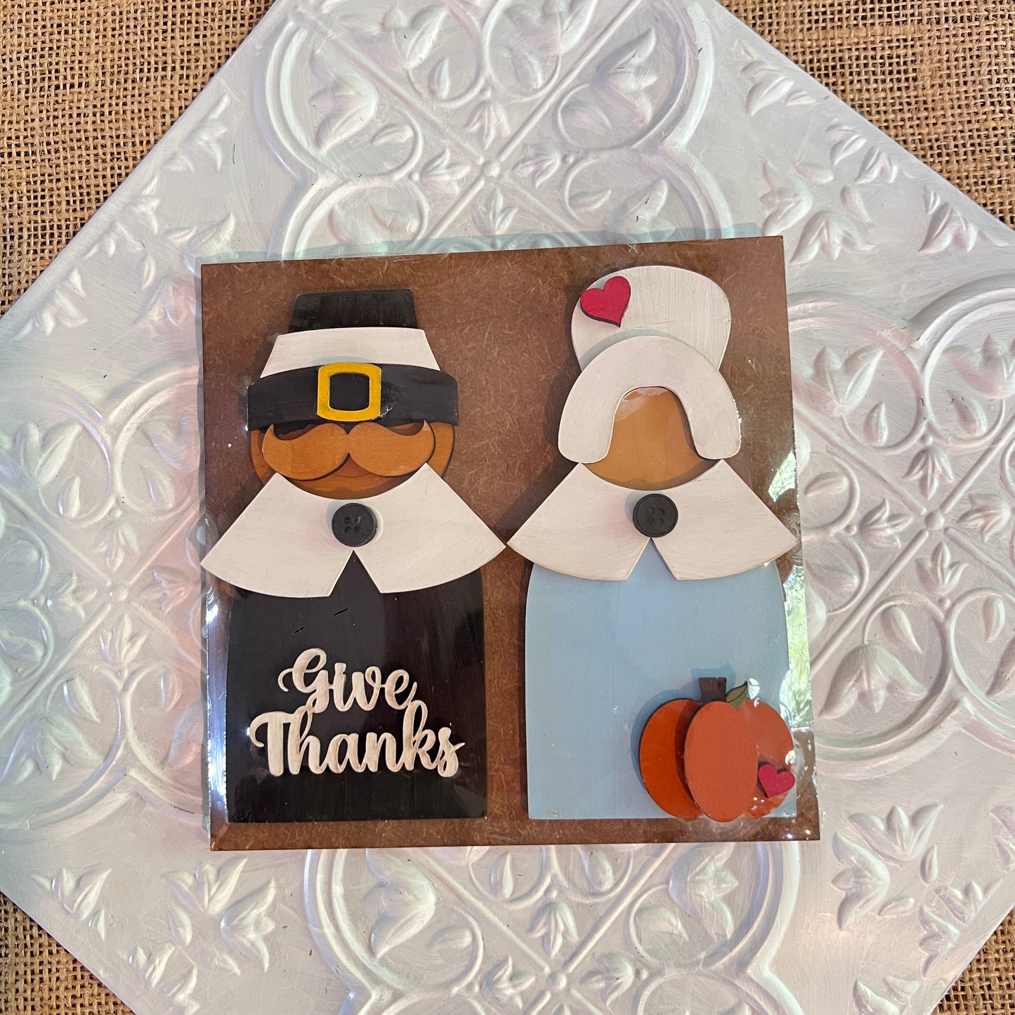 Janet's Craft Corner 6 inch squares feature wooden cutouts of a Pilgrim man and woman on a decorative tile, with “Give Thanks” on the man's outfit and a pumpkin by the woman, perfect for Thanksgiving decor.