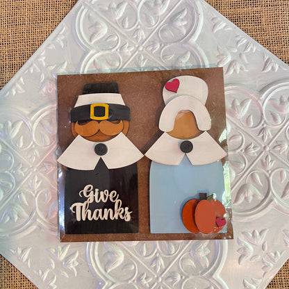 Janet's Craft Corner 6 inch squares feature wooden cutouts of a Pilgrim man and woman on a decorative tile, with “Give Thanks” on the man's outfit and a pumpkin by the woman, perfect for Thanksgiving decor.