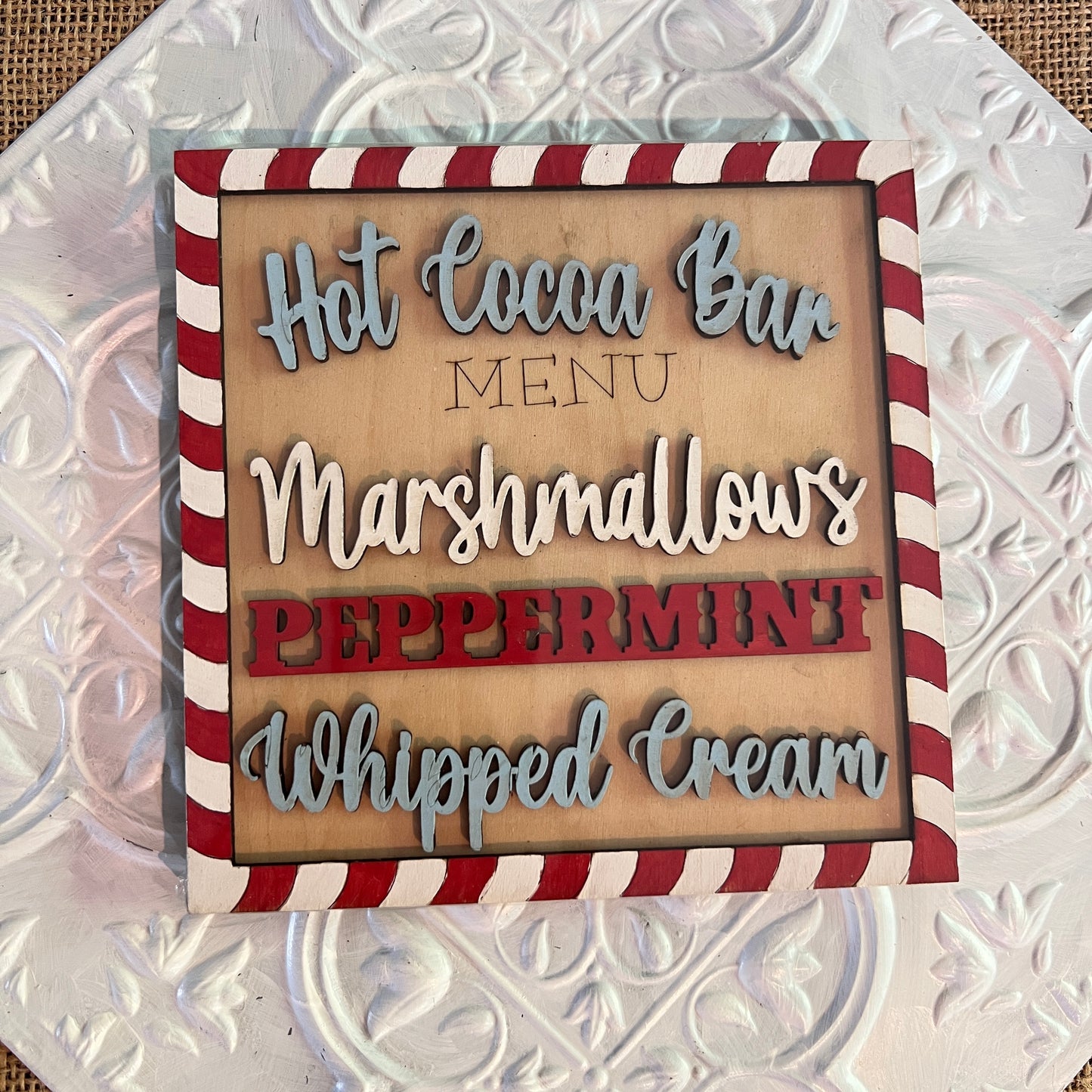 Janet's Craft Corner 6 inch squares: A wooden sign with a red and white candy cane striped border displays a hot cocoa bar menu—marshmallows, peppermint, and whipped cream—on a textured white background.