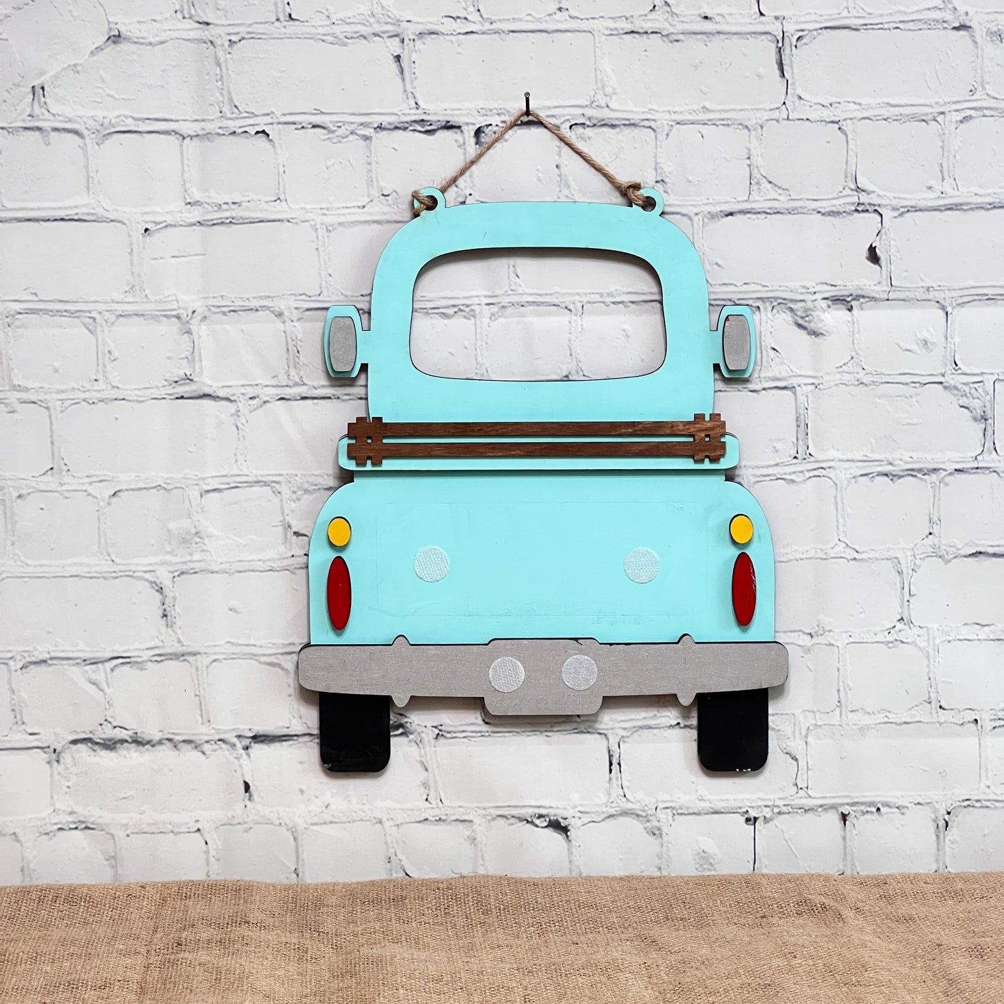 A light blue Antique Truck Interchangeable Base - Painted by Janet's Craft Corner hangs on a white brick wall above burlap. Seen from the back with wood accents and colorful tail lights, it's perfect for customizable décor.