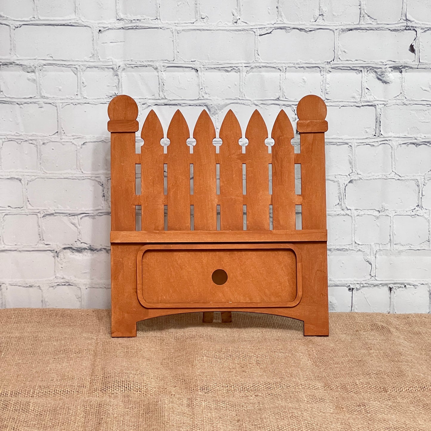 The Garden Fence Interchangeable Base - Painted by Janet's Craft Corner is a small wooden organizer with pointed slats, a bottom drawer, and sits on burlap by a white brick wall. Easily customized with interchangeable inserts for DIY home decor.