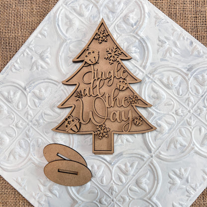 Introducing the "Standing Christmas Tree Shelf Sitters" by Janet's Craft Corner: This enchanting wooden decoration features "Jingle all the Way" intricately engraved, surrounded by beautifully carved bells and snowflakes. It's ideal for adding a festive touch to your holiday decor. Displayed on a textured white tile background, it creates a charming shelf display alongside two additional separate wooden pieces.