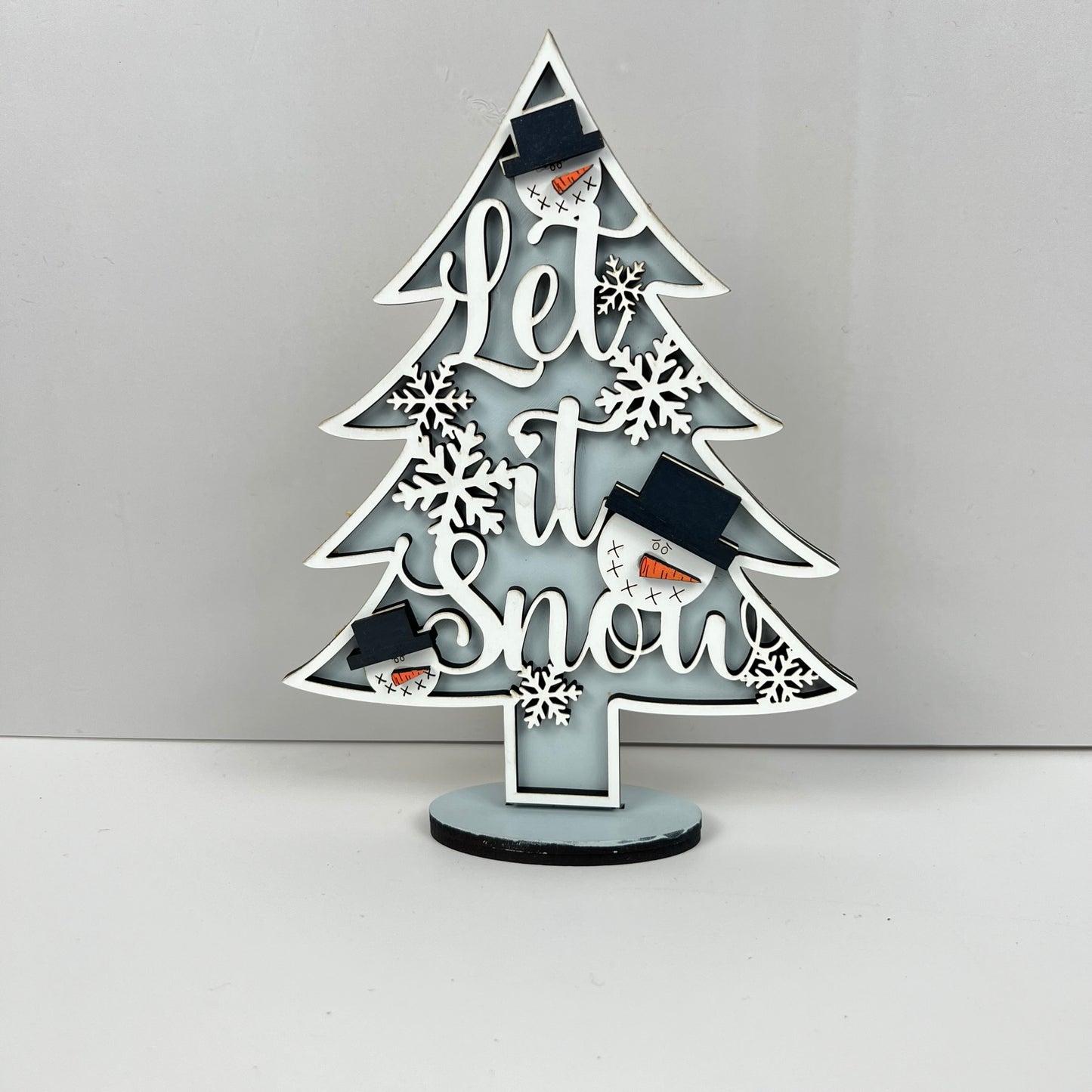 Janet's Craft Corner presents the Standing Christmas Tree Shelf Sitters, a beautifully decorative wooden tree ideal for holiday displays. It features "Let it Snow" intricately carved into the wood and is adorned with white snowflakes and adorable snowman faces sporting black hats. This festive decor stands elegantly on a minimalist dark base against a light backdrop, making it perfect for any seasonal shelf setting.