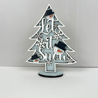 Janet's Craft Corner presents the Standing Christmas Tree Shelf Sitters, a beautifully decorative wooden tree ideal for holiday displays. It features "Let it Snow" intricately carved into the wood and is adorned with white snowflakes and adorable snowman faces sporting black hats. This festive decor stands elegantly on a minimalist dark base against a light backdrop, making it perfect for any seasonal shelf setting.