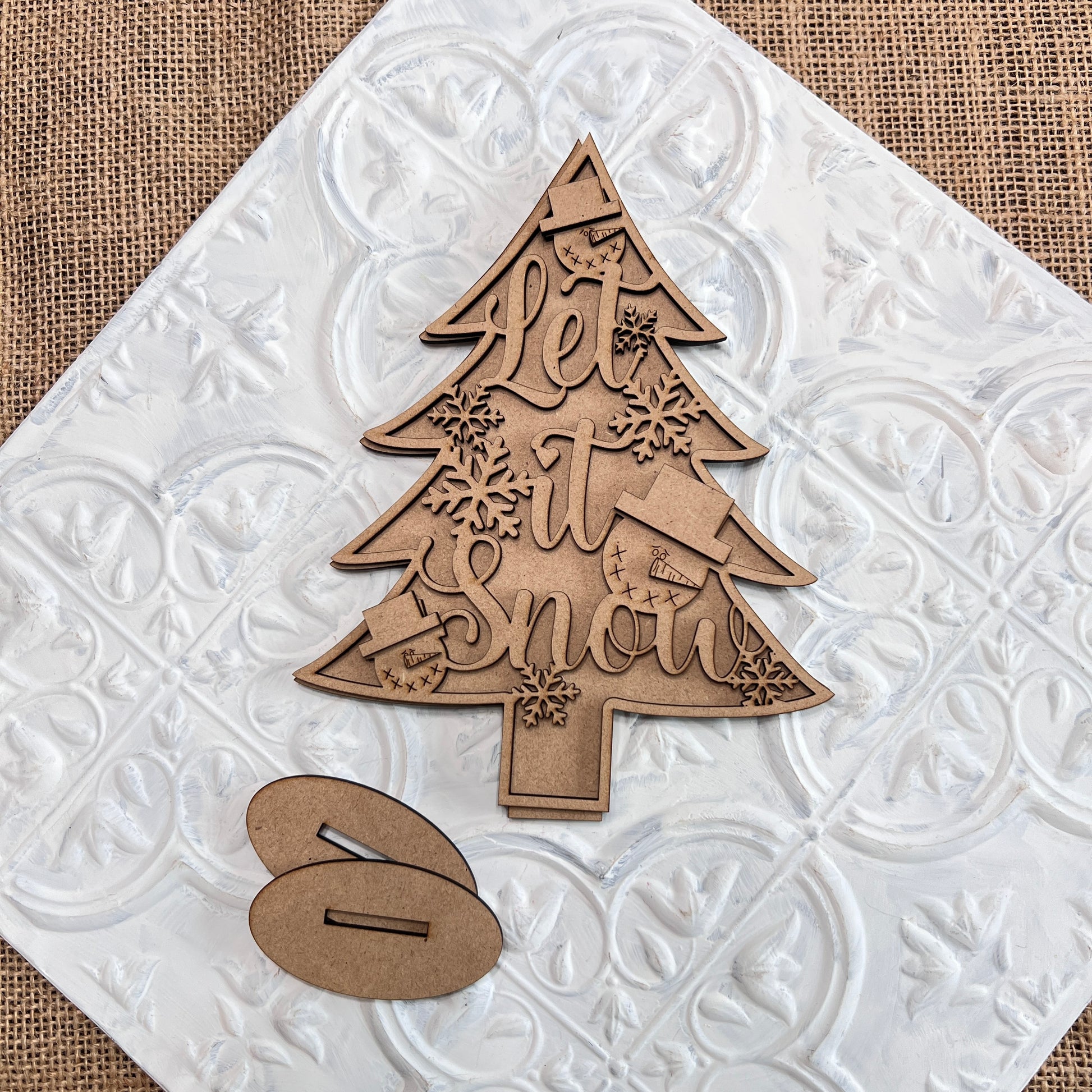 Janet's Craft Corner presents the Standing Christmas Tree Shelf Sitters, an exquisite wooden decoration ideal for holiday decor. It showcases "Let It Snow" engraved elegantly alongside snowflakes, mittens, and stockings. Placed on a textured white tile with a complementary oval stand piece beside it, this delightful item enhances any brown burlap backdrop with its charm.