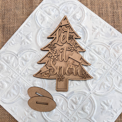 Janet's Craft Corner presents the Standing Christmas Tree Shelf Sitters, an exquisite wooden decoration ideal for holiday decor. It showcases "Let It Snow" engraved elegantly alongside snowflakes, mittens, and stockings. Placed on a textured white tile with a complementary oval stand piece beside it, this delightful item enhances any brown burlap backdrop with its charm.
