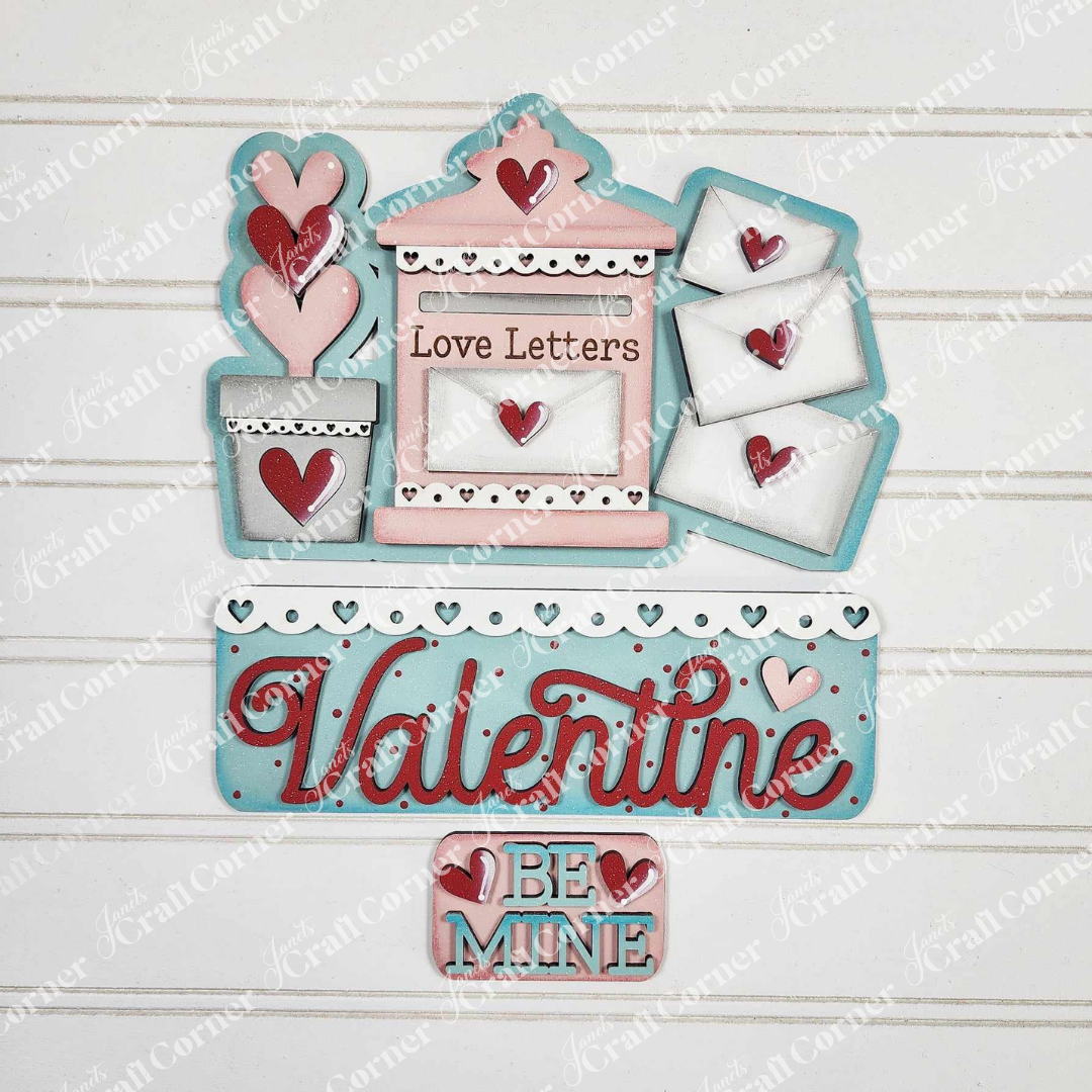 Janet's Craft Corner offers the Love Letters Interchangeable Insert: a Valentine's craft set featuring a pink and blue mailbox labeled "Love Letters," three envelopes with heart seals, and wooden heart cutouts in a flower pot adorned with "Valentine" and "Be Mine" on a white background.