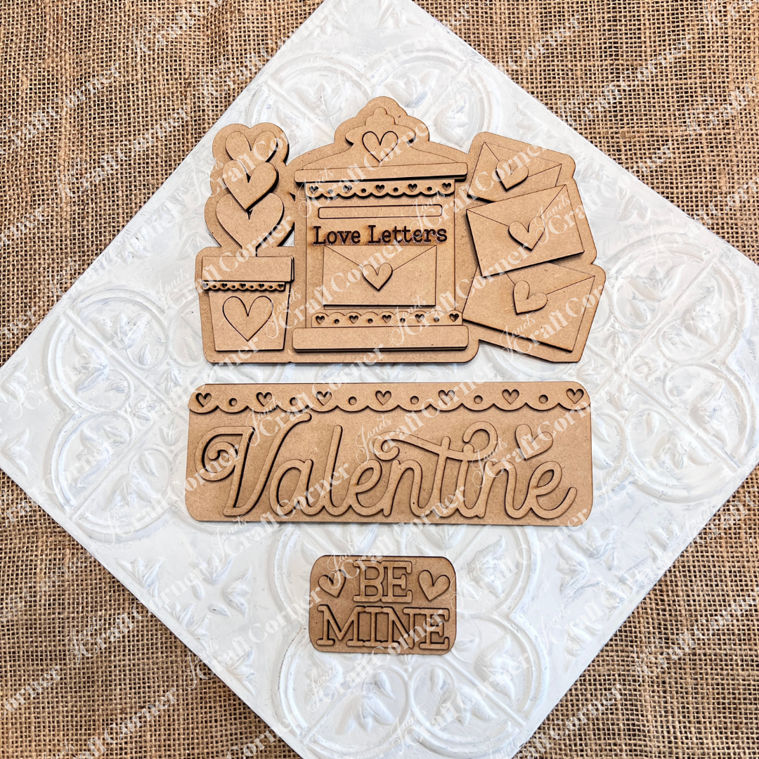 Janet's Craft Corner presents the Love Letters Interchangeable Insert: hand-painted wooden Valentine's cutouts including a "Love Letters" mail slot, a "Valentine" sign, and a "Be Mine" plaque with heart details on burlap over a textured white background.