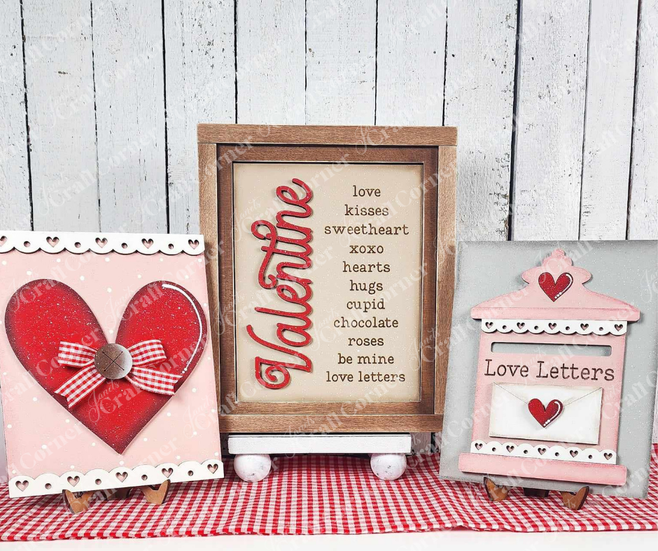 The Love Letters Mailbox Interchangeable Sign kit from Janet's Craft Corner features three charming wooden signs: a red heart, love-themed words "Valentine," and a pink mailbox labeled "Love Letters." Set against a red gingham cloth on a white wooden backdrop, it's perfect for interchangeable home decor.