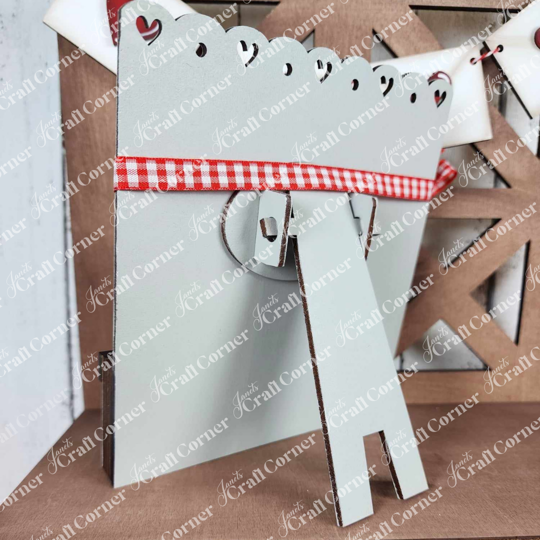 The Valentine Love Letters Decor Set from Janet's Craft Corner features a light gray stand with heart cut-out details, a personalized charm, and a red and white gingham ribbon, perfect for adding warmth to your home's wooden backdrop.