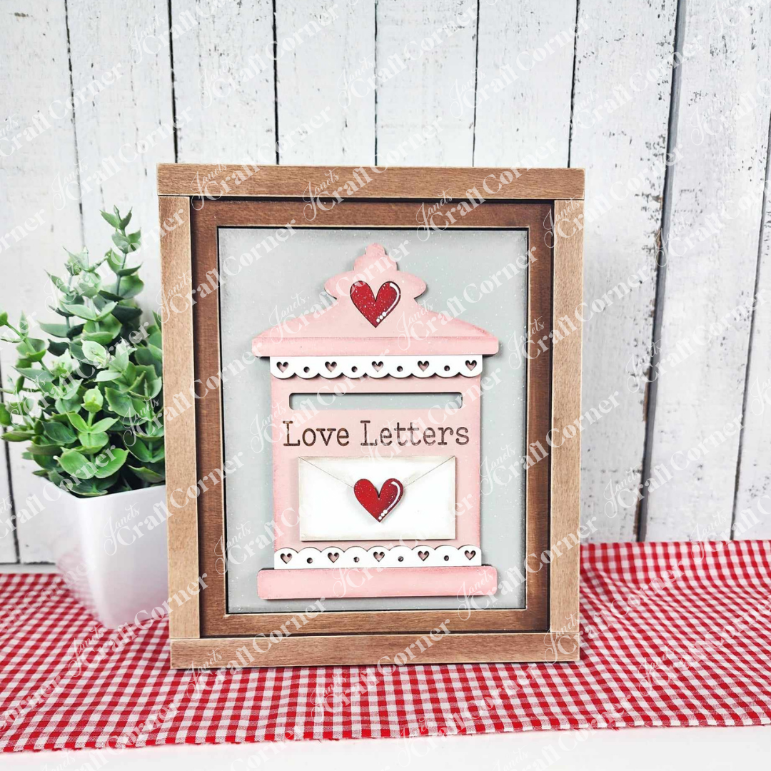 Janet's Craft Corner offers the "Love Letters Mailbox" interchangeable sign kit, a framed decorative piece showing a pink mailbox with hearts and "Love Letters" text. The scene includes a red and white checkered cloth, potted plant, and wooden wall backdrop. Ideal for seasonal home decor.