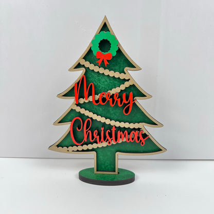Add festive charm to your shelf with the Heartland Standing Christmas Tree Shelf Sitter—painted green, adorned with gold garland, a wreath with a red bow on top, and "Merry Christmas" in red script. Perfect holiday decor!.