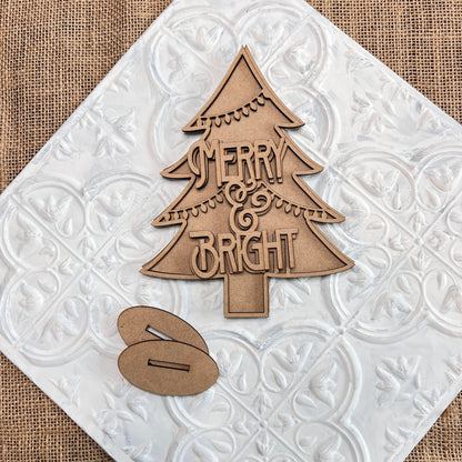 Presenting the *Standing Christmas Tree Shelf Sitters* from Janet's Craft Corner: a wooden cutout of a standing Christmas tree on a decorative white tile background, featuring the festive words "Merry & Bright." This charming piece is perfect for your holiday decor and includes two small, oval-shaped wooden accents beneath the tree for an elegant shelf display.