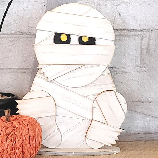 The Mummy Shelf Sitter DIY home decor kit by Janet's Craft Corner features a cartoon-style figure with yellow eyes wrapped in white bandages, perfect for positioning next to a small orange pumpkin. Enthusiasts of hand-painted DIY projects will appreciate using it to create a charming display against a white brick background.