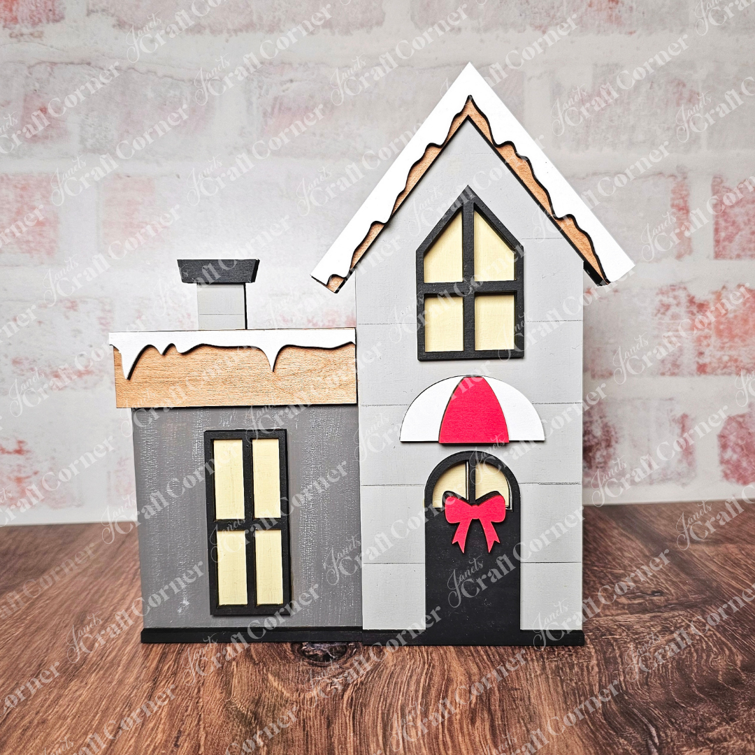 This delightful piece from Janet's Craft Corner's Christmas Village Shelf Sitters - Set of 4 features a decorative wooden house with a gray chimney and snow-covered roof. It showcases a charming design, featuring a door embellished with a red bow under a red and white awning, set against its wooden surface and brick background.