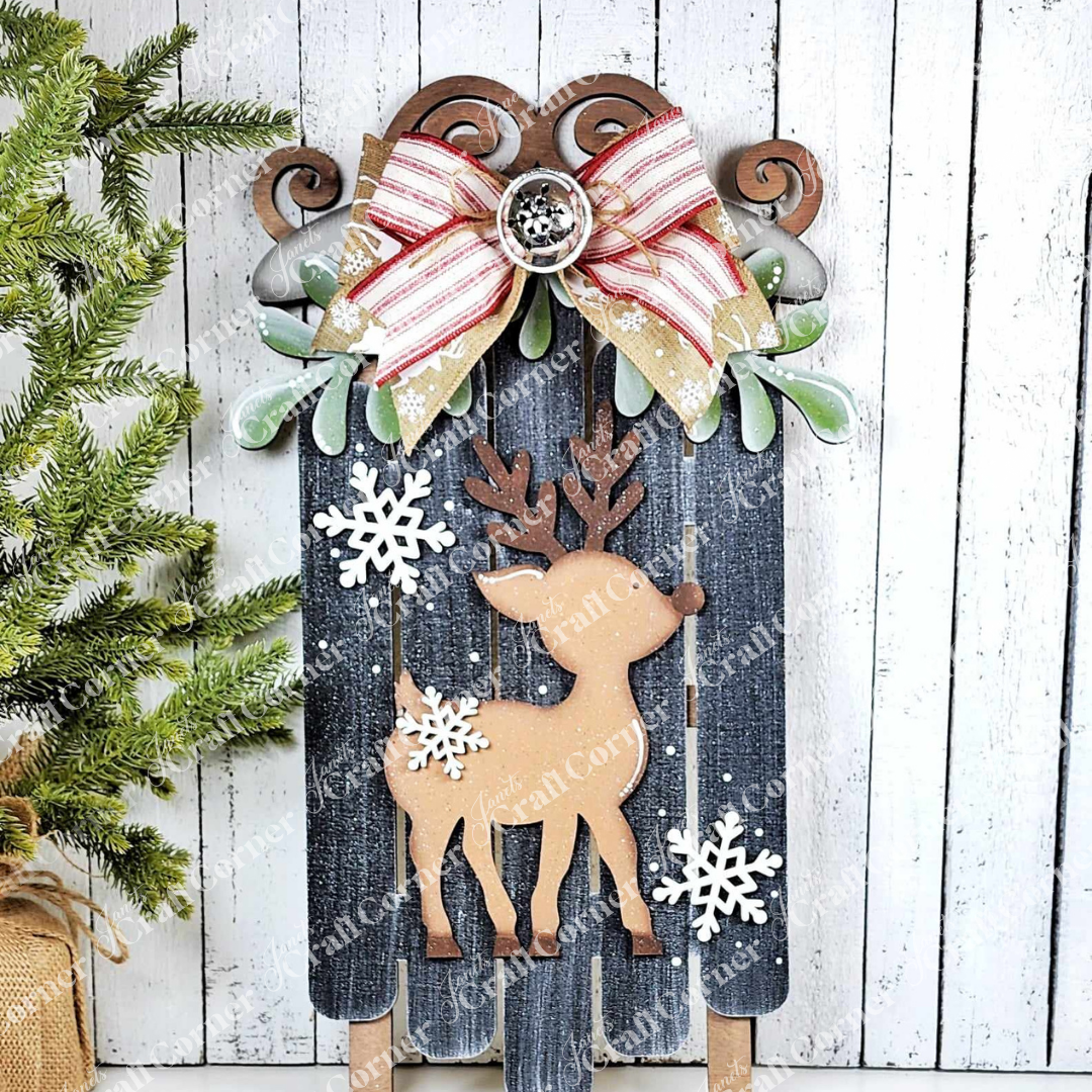 The Oh Deer Sled by Janet's Craft Corner is a delightful hand-painted wooden Christmas decoration showcasing an enchanting reindeer adorned with snowflakes. It’s beautifully finished with a red and white striped bow accompanied by green leaves. Ideal for enhancing home décor, it can be elegantly displayed against a white wooden wall with a petite evergreen tree on the left.