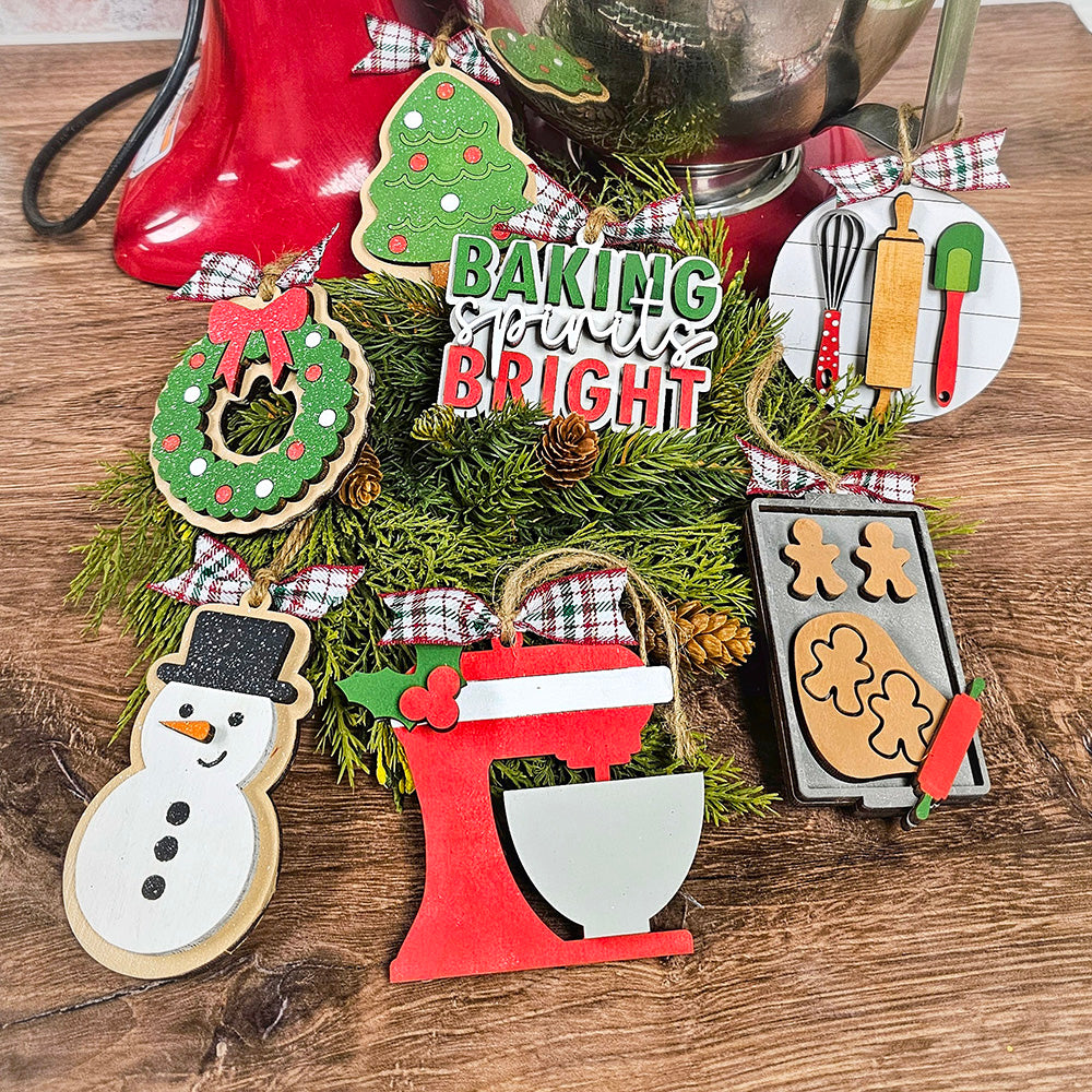 A festive display featuring Christmas-themed ornaments from Janet's Craft Corner showcases the DIY delights of their Sugar Cookie Ornaments - Set of 3. The decorations, including a wreath, snowman, mixer, gingerbread cookies, baking set, and tree, are elegantly adorned with plaid ribbon bows and greenery on a wooden surface.