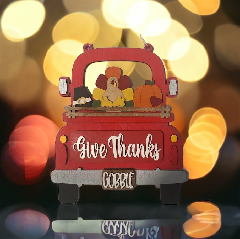 The DIY home decor kit from Janet's Craft Corner, the "Give Thanks Interchangeable Insert," includes a charming decorative red truck with "Give Thanks" and "Gobble" text. It features interchangeable inserts such as a small turkey and pumpkins, all set against a warm backdrop of defocused bokeh lights that enhance the festive Thanksgiving ambiance.