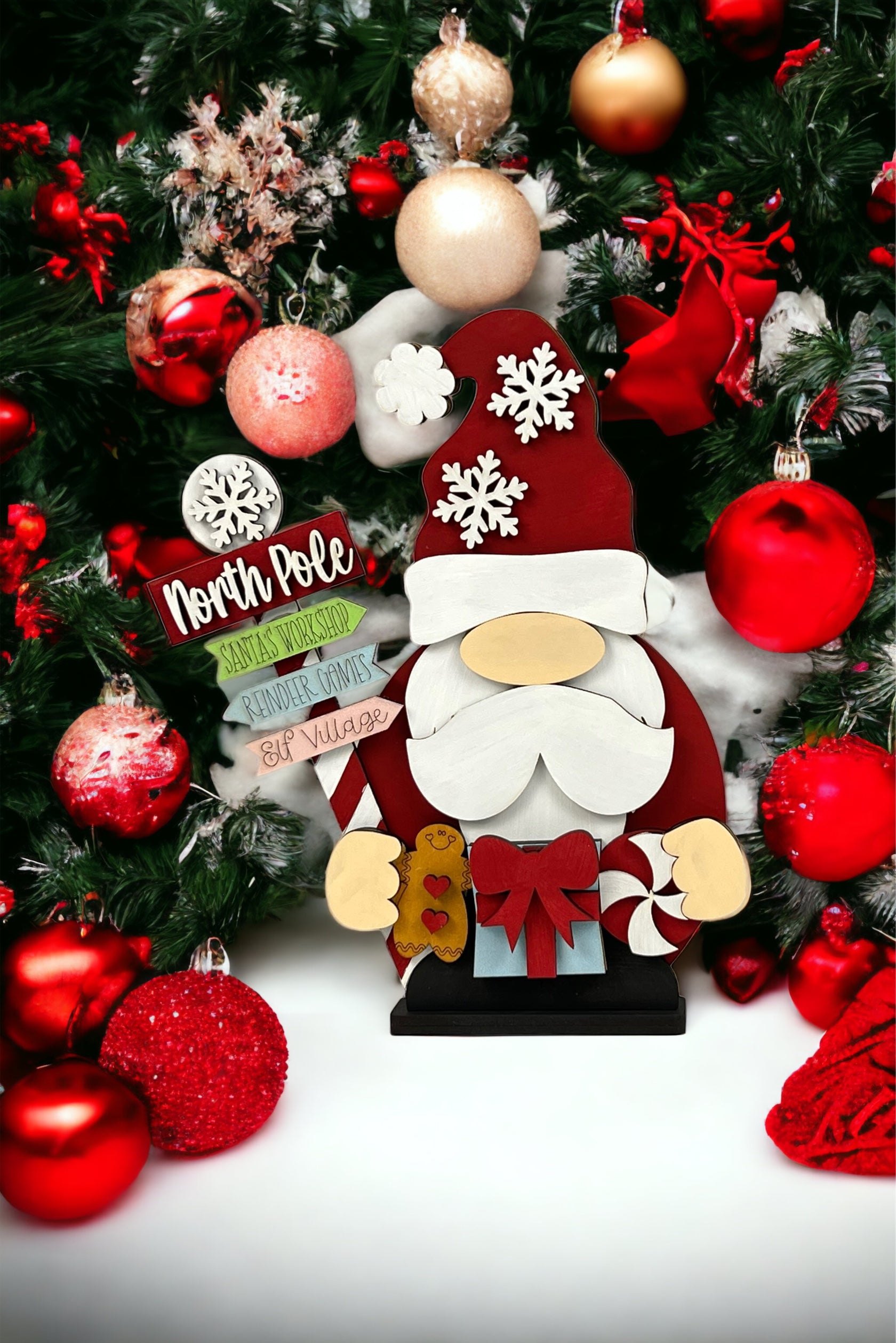 Transform your holiday decor into a festive scene with Janet's Craft Corner’s Santa Gnome Shelf Sitter from their DIY holiday decor craft kit. This delightful decoration features a Santa gnome holding candy canes and a present, surrounded by vibrant red and white ornaments and lush green pine branches. Complete the look with a charming wooden sign pointing to whimsical North Pole destinations like Santa’s Workshop and Elf Village, bringing your holiday imagination to life.