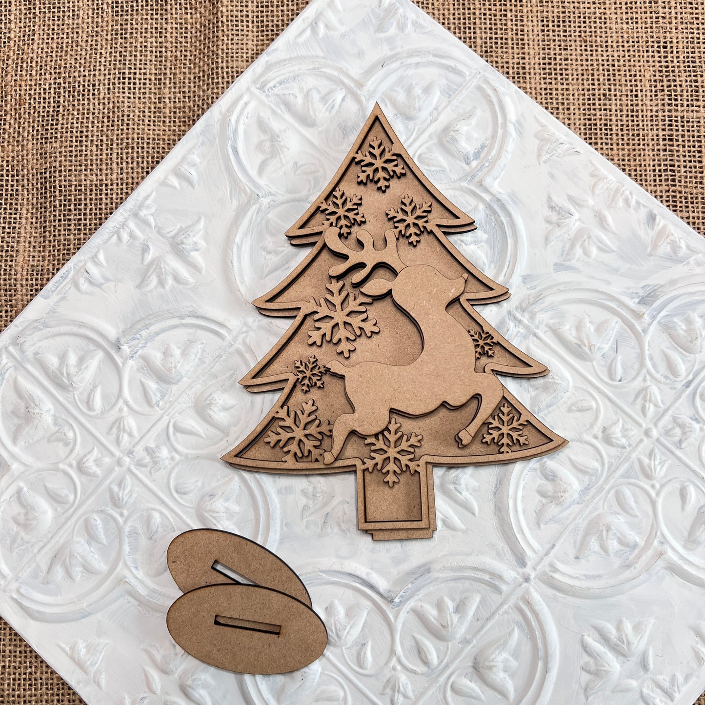 The Standing Christmas Tree Shelf Sitters by Janet's Craft Corner showcase a laser-cut wooden Christmas tree ornament adorned with a reindeer and snowflake design, elegantly displayed on an ornate white tile against a burlap backdrop. Perfect for holiday decor, two oval wooden bases are artfully arranged in the foreground.