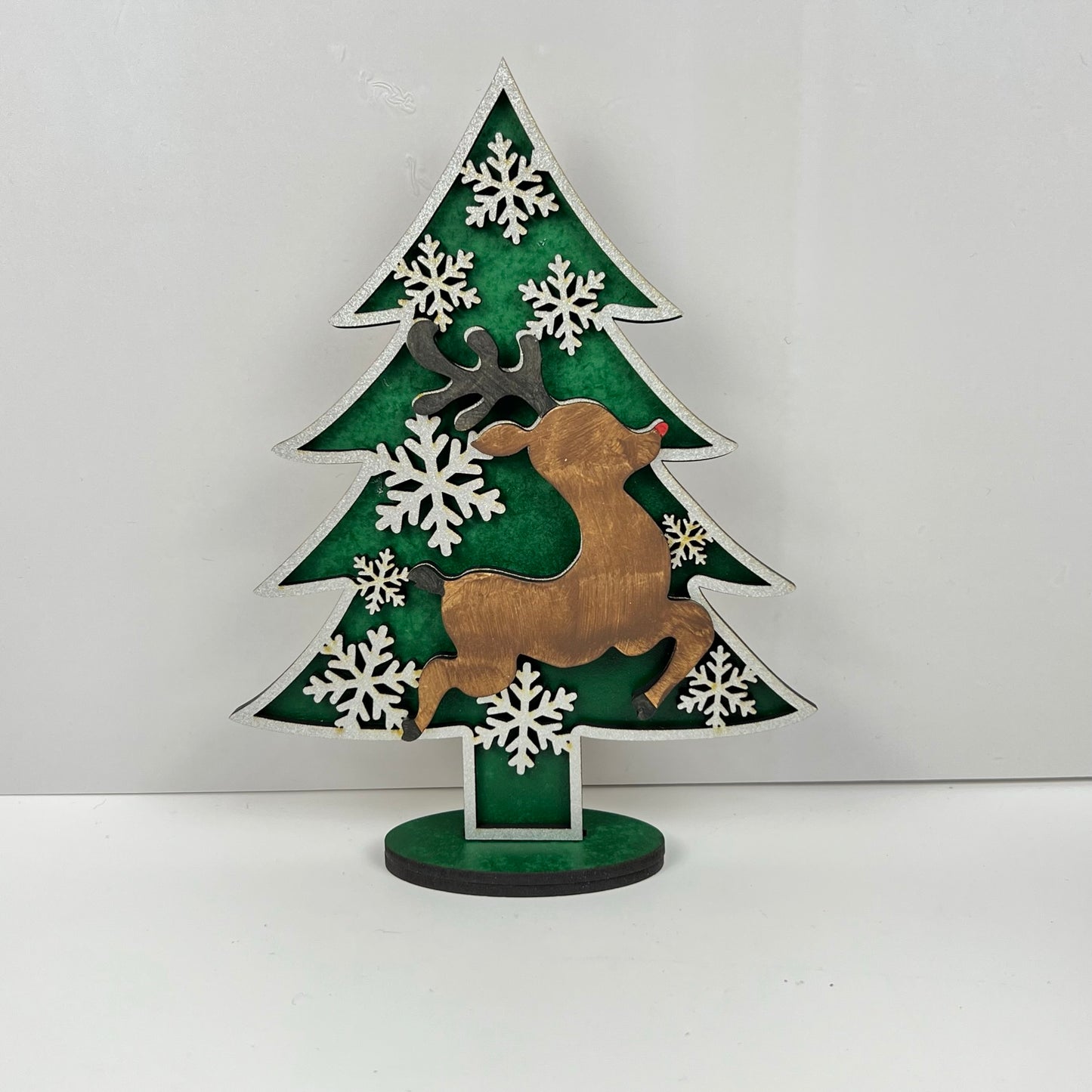 Introducing the Standing Christmas Tree Shelf Sitters by Janet's Craft Corner, a decorative piece featuring a wooden tree design with a bronze reindeer silhouette and white snowflakes set against a green background. This elegant holiday decor stands gracefully on a round base, making it an ideal addition to your festive decorations.