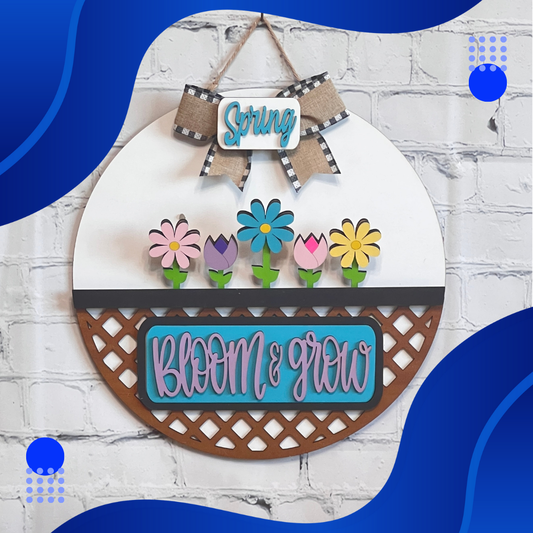 On a white brick wall hangs Janet's Craft Corner's "Bloom & Grow Interchangeable Insert," a round sign from their DIY decor kit. It features colorful flowers with "Spring Blooms" above and "Bloom & Grow" below, set against blue wave-like shapes and dotted accents, echoing vintage truck curves.
