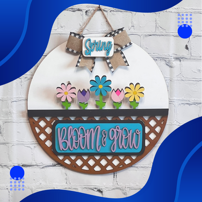 On a white brick wall hangs Janet's Craft Corner's "Bloom & Grow Interchangeable Insert," a round sign from their DIY decor kit. It features colorful flowers with "Spring Blooms" above and "Bloom & Grow" below, set against blue wave-like shapes and dotted accents, echoing vintage truck curves.